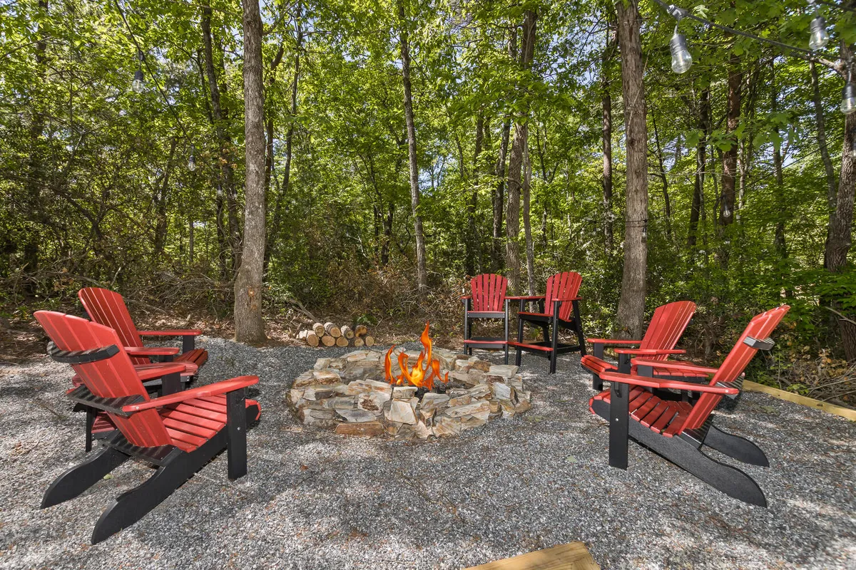 The exterior fire pit area is shown ringed by red Adirondack chairs, set amidst tall trees. The fire pit is the centerpiece, with the setting suggesting communal gatherings, storytelling, and laughter under the stars. The mood is relaxed and inviting.