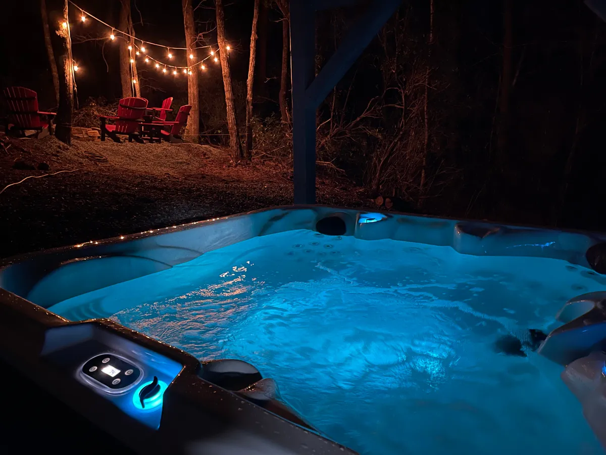 This exterior image features a hot tub with softly illuminated water, string lights overhead, and a cluster of red Adirondack chairs arranged around a fire pit. The setting exudes a cozy, inviting atmosphere perfect for stargazing, relaxing soaks, or sharing stories by the fire.