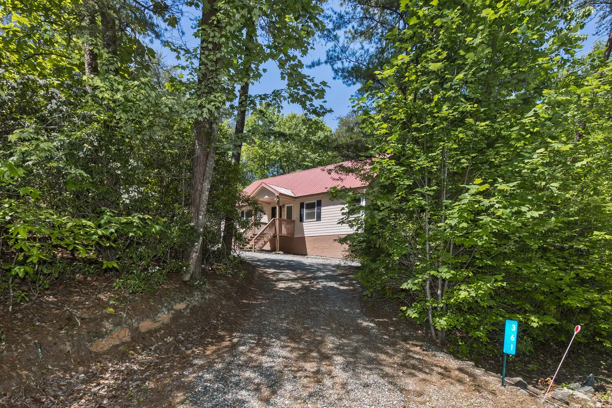 A peaceful exterior view of the house with its distinctive red roof, surrounded by tall trees and greenery. The gravel driveway leads up to the home, and the natural forested setting creates a secluded, inviting atmosphere.