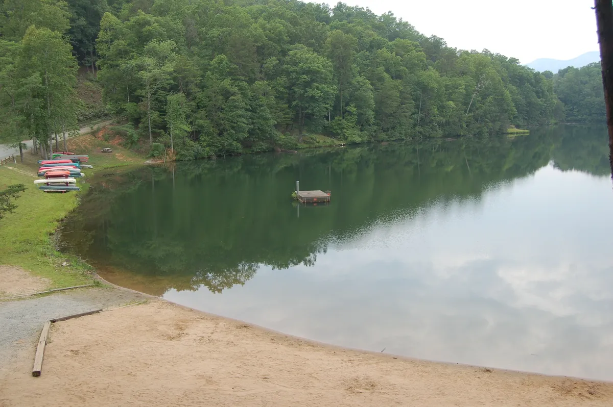 This image features a calm, pristine lake surrounded by lush trees, with a wooden dock and kayaks visible. The scene evokes a tranquil, private setting perfect for swimming, boating, fishing, and enjoying the natural beauty of Riverbend's exclusive amenities.