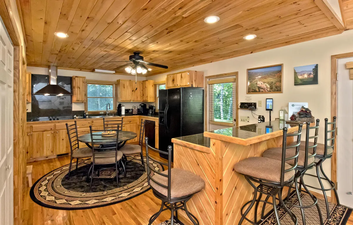 This image features a warm, inviting kitchen with wooden cabinetry, modern black appliances, a round dining table, and bar stools. The space is clearly designed for gathering and culinary activities, supporting both cooking and casual dining. The setup encourages socializing and interaction between the kitchen and dining area.