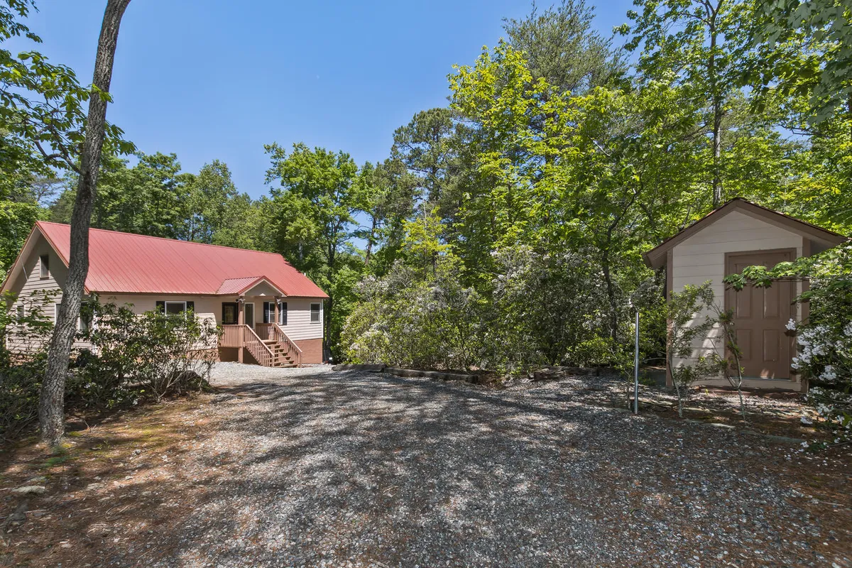 An inviting exterior shot of the house with its red roof, gravel driveway, and surrounding trees. The image includes a glimpse of a storage shed and abundant nature, emphasizing privacy and ample space for exploration.