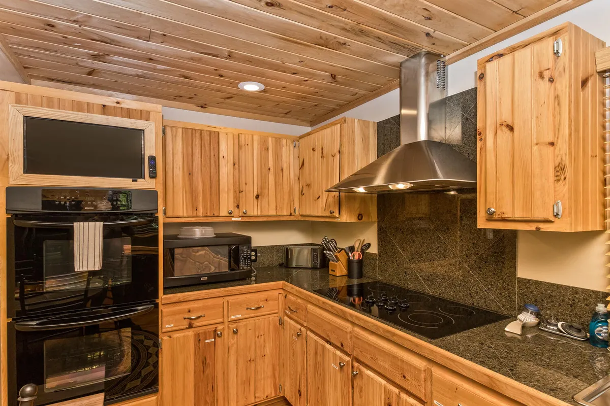 This image spotlights the kitchen's modern finishes, including wood cabinetry, a granite countertop, stainless steel appliances (notably a double oven and vent hood), and a functional yet cozy workspace. The setup is ideal for group meal preparation and entertaining.
