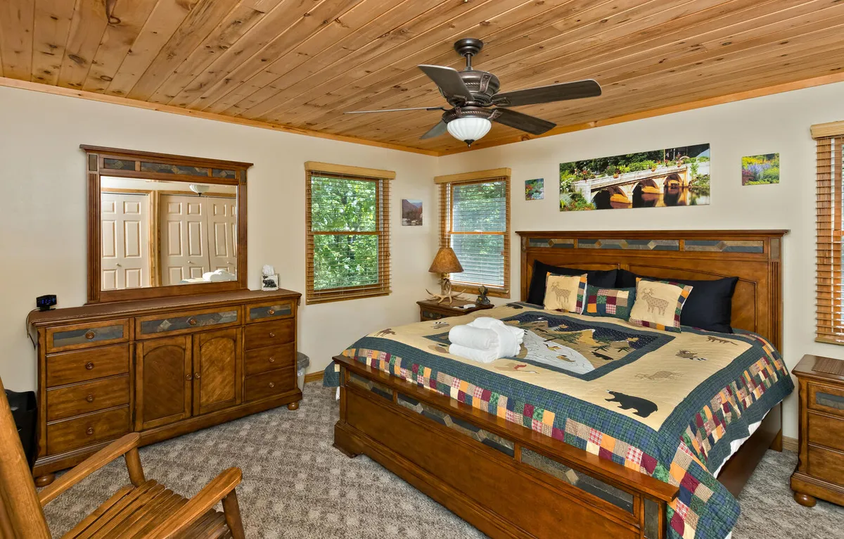 A welcoming bedroom with a wooden bed frame, a colorful quilt, a dresser with a mirror, and windows that allow natural light to pour in. The scene is cozy and bright, offering a restful atmosphere ideal for relaxation and sleep.