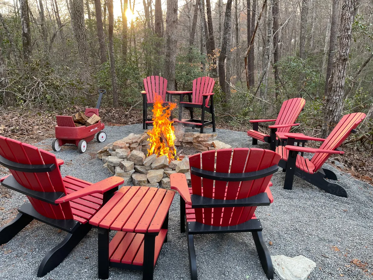 The exterior of a luxury cabin set against a peaceful wooded backdrop, featuring a crackling fire pit surrounded by vibrant red Adirondack chairs and a rustic wagon nearby. The scene invites relaxation and connection as the sun sets, promising cozy evenings and cherished memories.