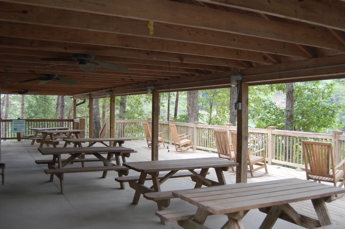 A casual outdoor space featuring wooden picnic tables and classic rocking chairs arranged on a deck with wooden railings. The area invites guests to relax, dine, and enjoy the natural surroundings, with greenery visible beyond the deck.
