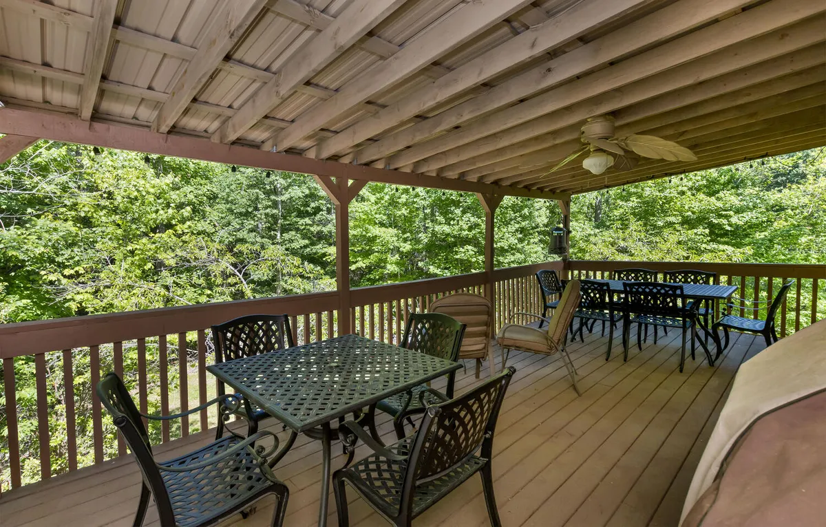 This image captures a tranquil exterior deck area, featuring a dining table and seating arrangement surrounded by lush greenery. The space is shaded by a ceiling fan, offering a comfortable setting for outdoor dining or relaxation. The deck overlooks the natural landscape, providing a seamless connection between indoor comfort and outdoor adventure.