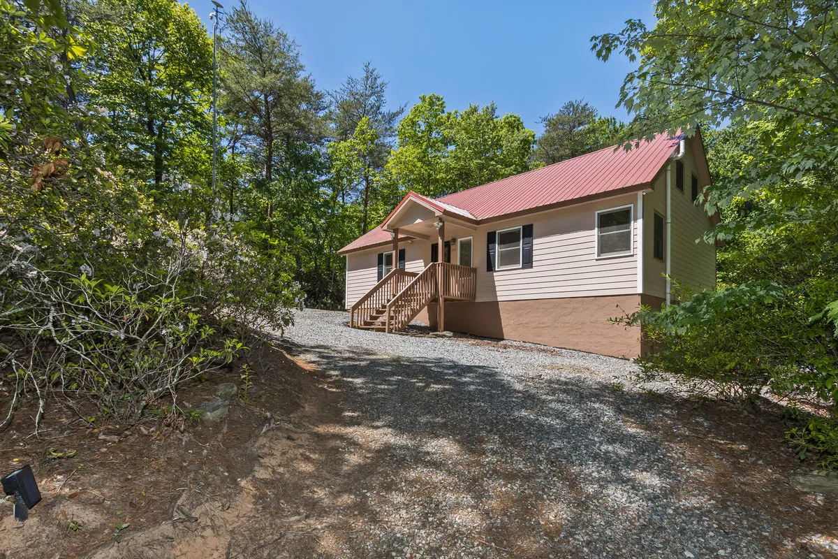 This image captures the house’s facade, front porch, gravel driveway, and the lush trees surrounding the home, all bathed in natural light. The scene highlights the peaceful mountain setting while showing the welcoming approach to the property, connecting the experience of arrival with the outdoors.