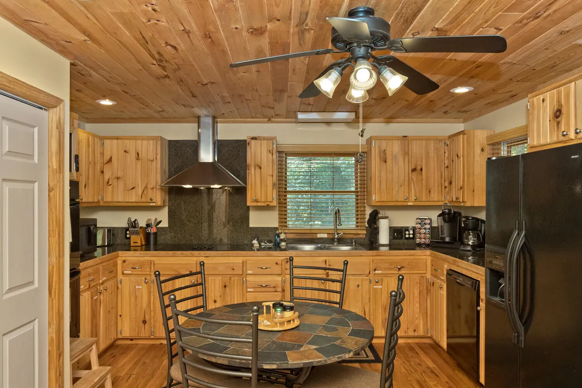 This photo presents another angle on the kitchen and dining space, highlighting wooden cabinets, black appliances, a round mosaic-topped table, and a ceiling fan. The scene is set for family gatherings and shared meals, with a warm, welcoming ambiance.