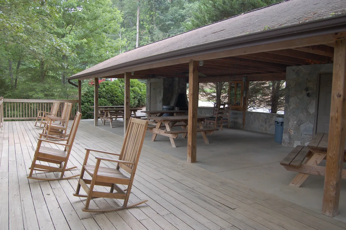 A charming exterior space featuring classic rocking chairs and wooden picnic tables set beside a rustic stone wall, surrounded by greenery. The communal setup encourages relaxation and gathering in a safe outdoor environment.